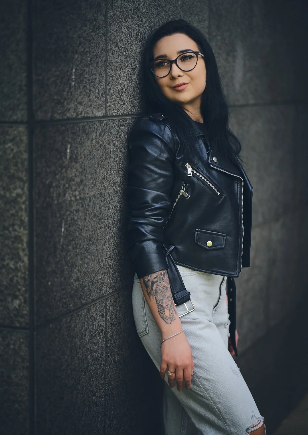 A woman with black hair and glasses leaning against a dark wall, wearing a black leather jacket, light-colored jeans, and tattoos on her left arm.