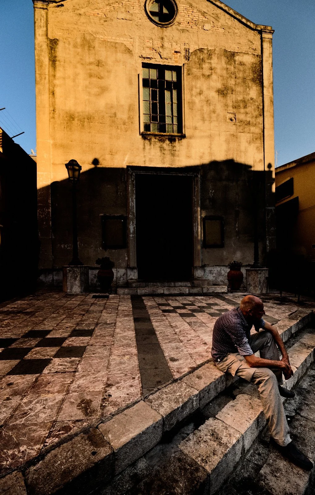 Old man sitting alone on marble steps before a weathered church facade half in shadow, Taormina, Sicily — Provincia Malinconica by Tom Imio