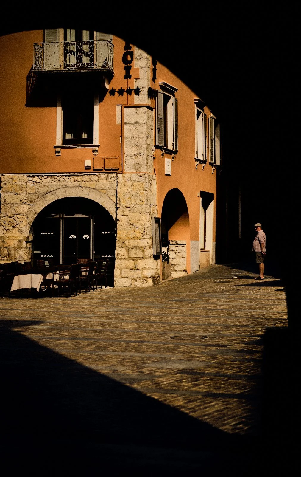 Solitary man standing in a sharp diagonal shadow beside a sunlit terracotta building with wrought-iron balcony in Riva del Garda, Italy — Provincia Malinconica by Tom Imio