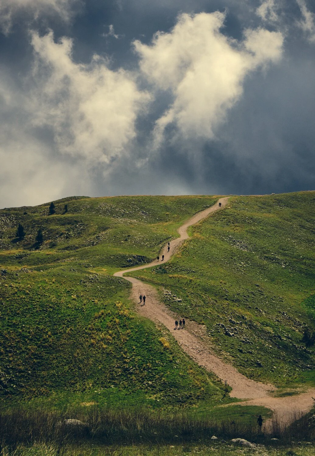 Hikers walking along a winding dirt trail across a green hillside under a cloudy sky.