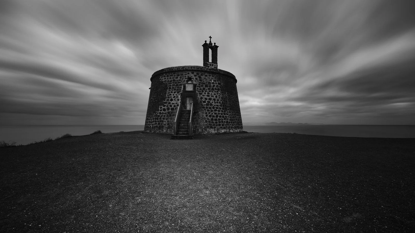 Black and white photo of a small, round stone chapel on a cliff, with steps leading up to the entrance, overlooking the ocean under a cloudy sky.