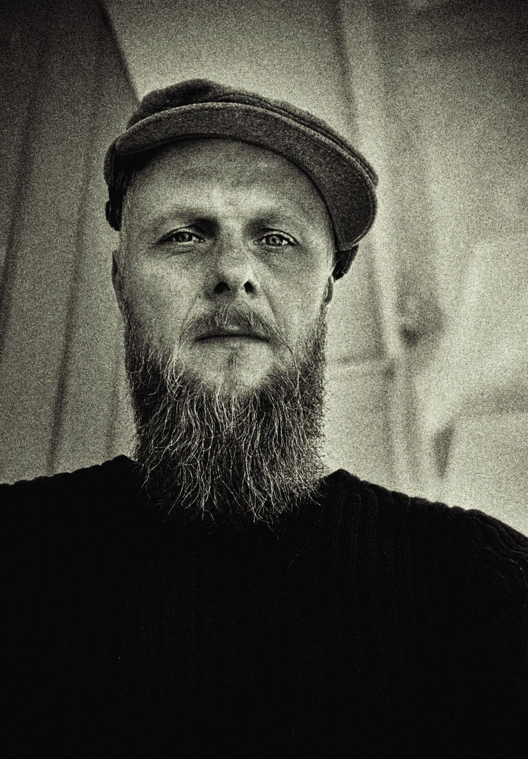 A black and white portrait of a man with a beard and mustache, wearing a cap and a dark shirt, looking directly at the camera.