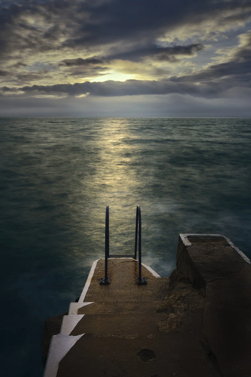 A view of a staircase descending into the ocean with a cloudy sky and the sun partially visible on the horizon.