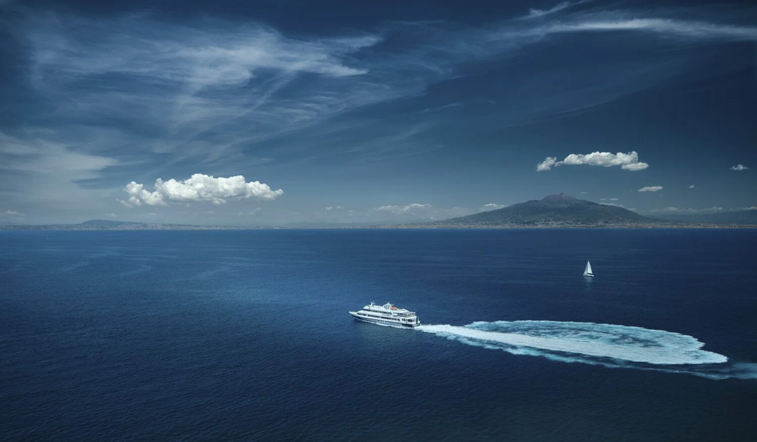 A large white yacht moving at high speed in the ocean creating a wake, with a sailboat in the distance, and an island with a mountain in the background under a partly cloudy sky.
