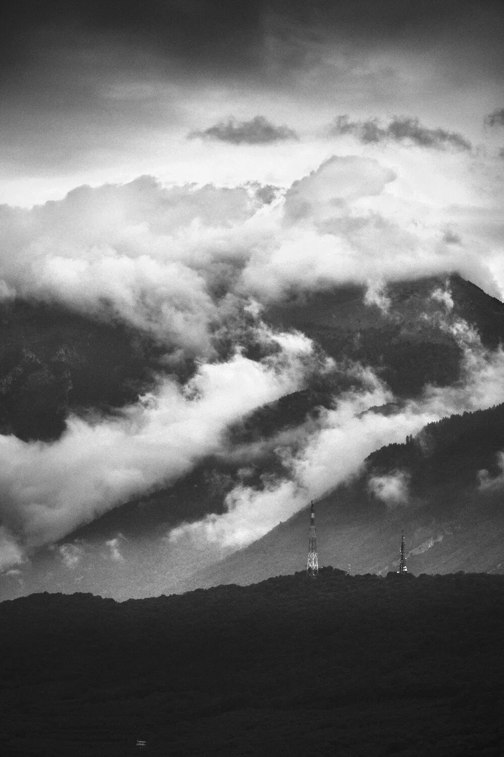 Black and white photograph of a mountain landscape with clouds and fog partially covering the mountains and two communication towers on a hill in the foreground.
