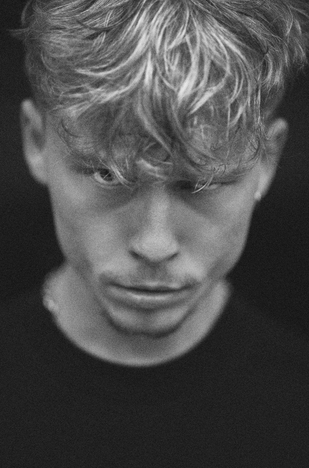 Close-up black and white portrait of a young man with tousled light hair looking down, half his face emerging from darkness — Echoes of Self by Tom Imio