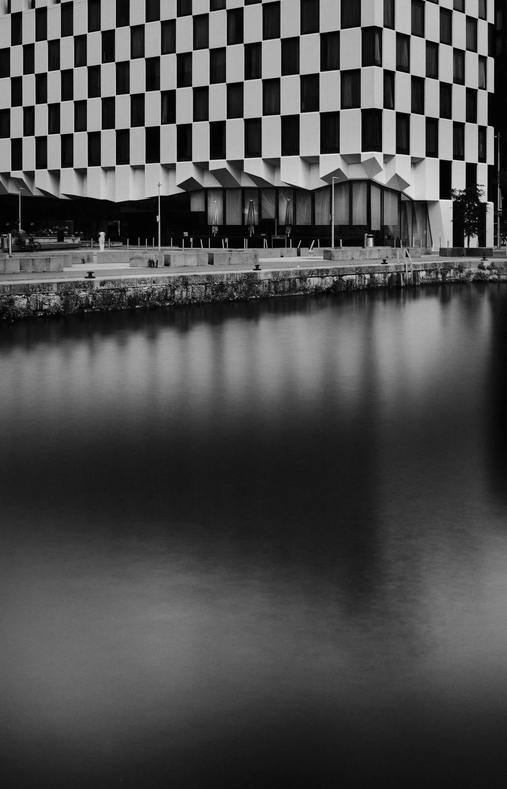 Modern building with a checkered facade, located by a body of water, with outdoor seating and streetlights in the foreground.