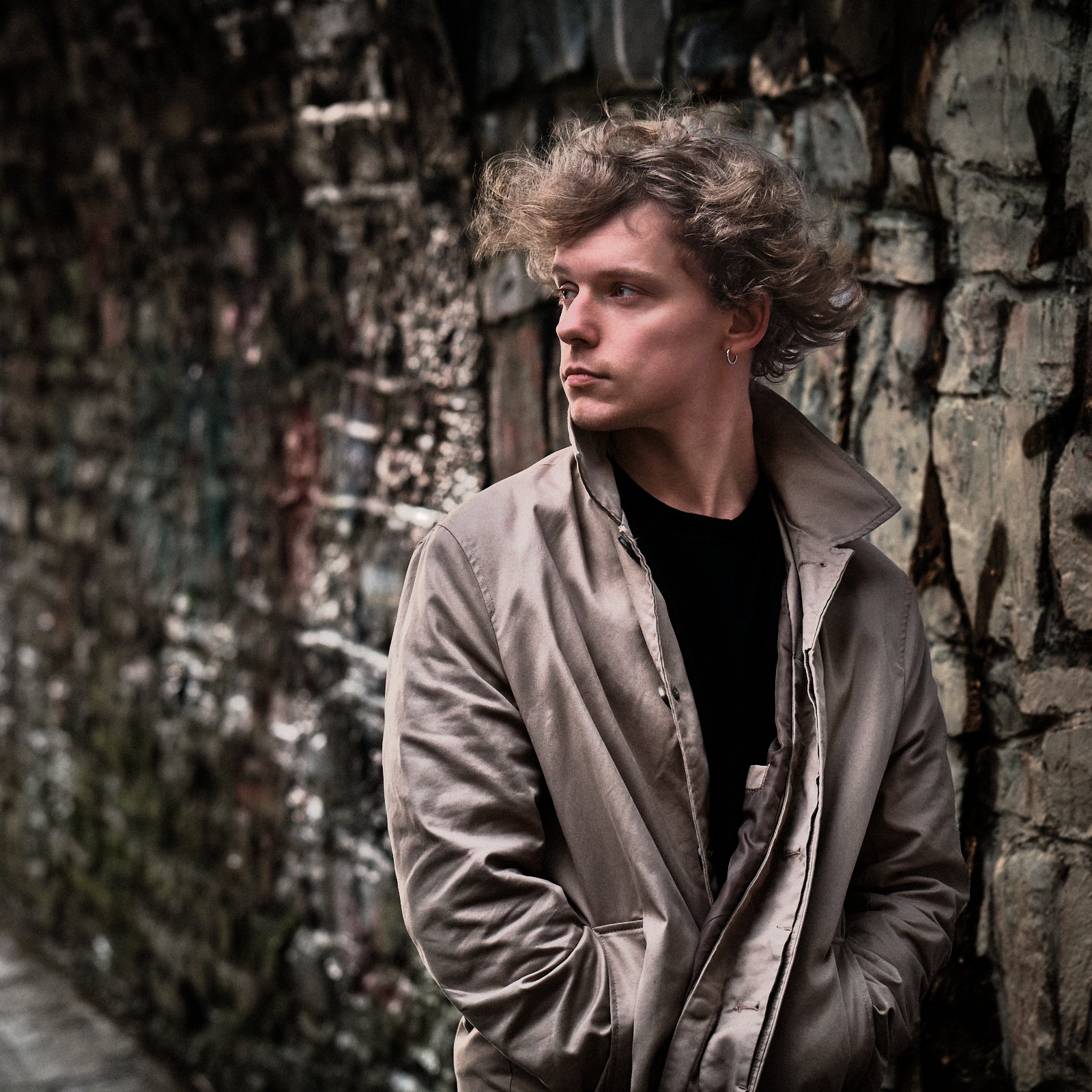 A young man with curly hair and earrings stands against a stone wall, looking to the side with a contemplative expression.