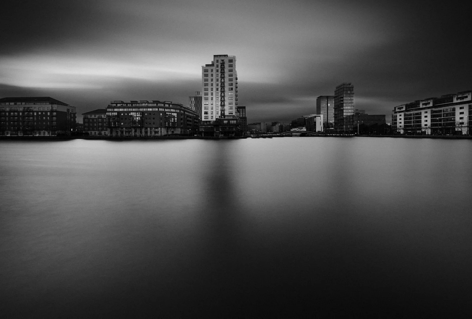 Black and white photo of a city skyline with tall buildings across a body of water, under a cloudy sky.