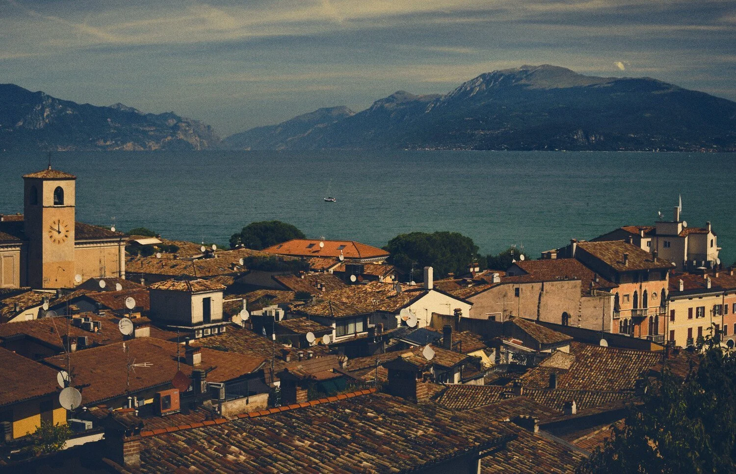 View of a coastal town with tiled rooftops, a clock tower, and satellite dishes, overlooking a lake with mountains in the background.