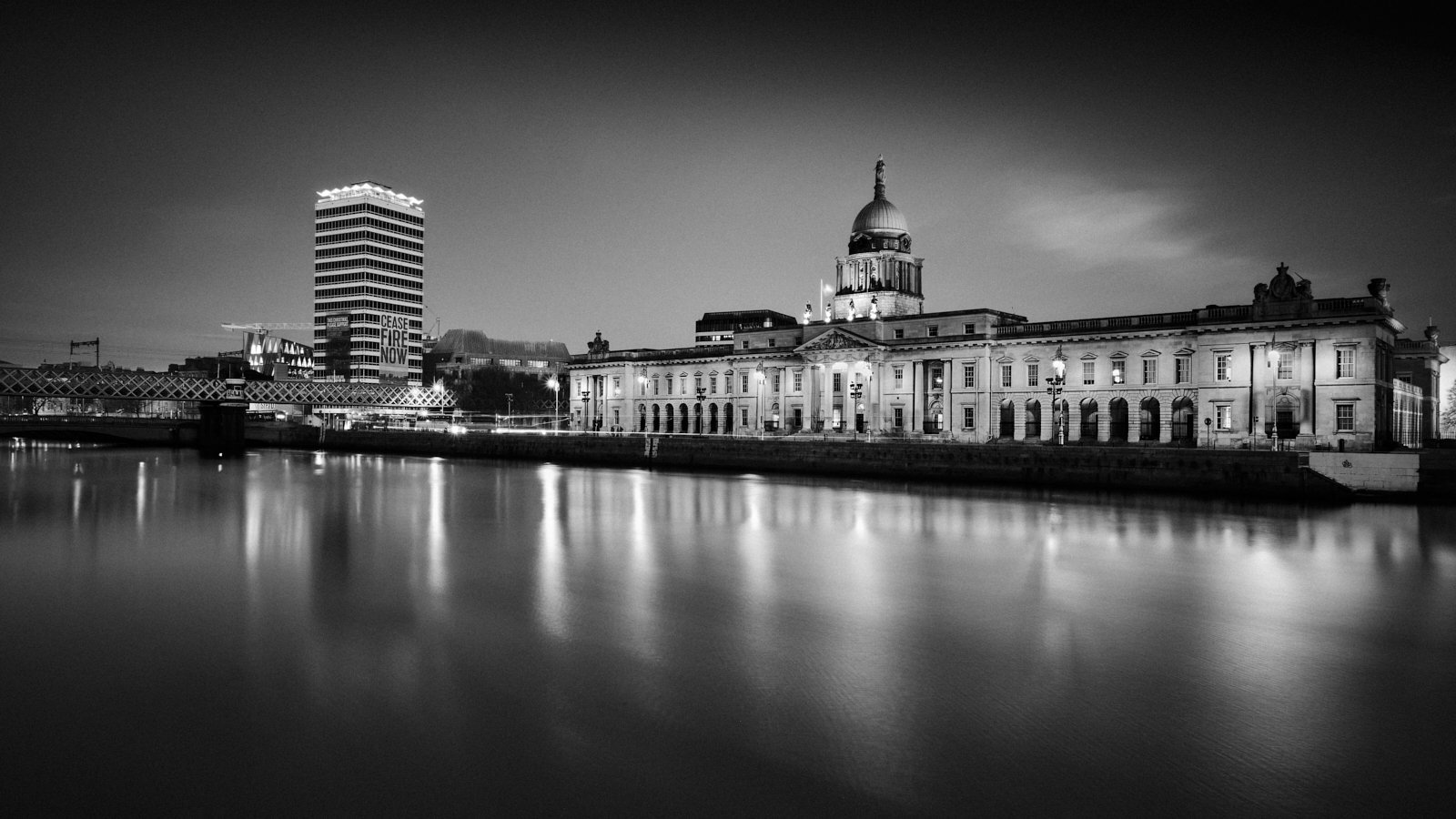 Black and white night view of a historic building with a dome next to a modern high-rise, reflecting in a calm river.