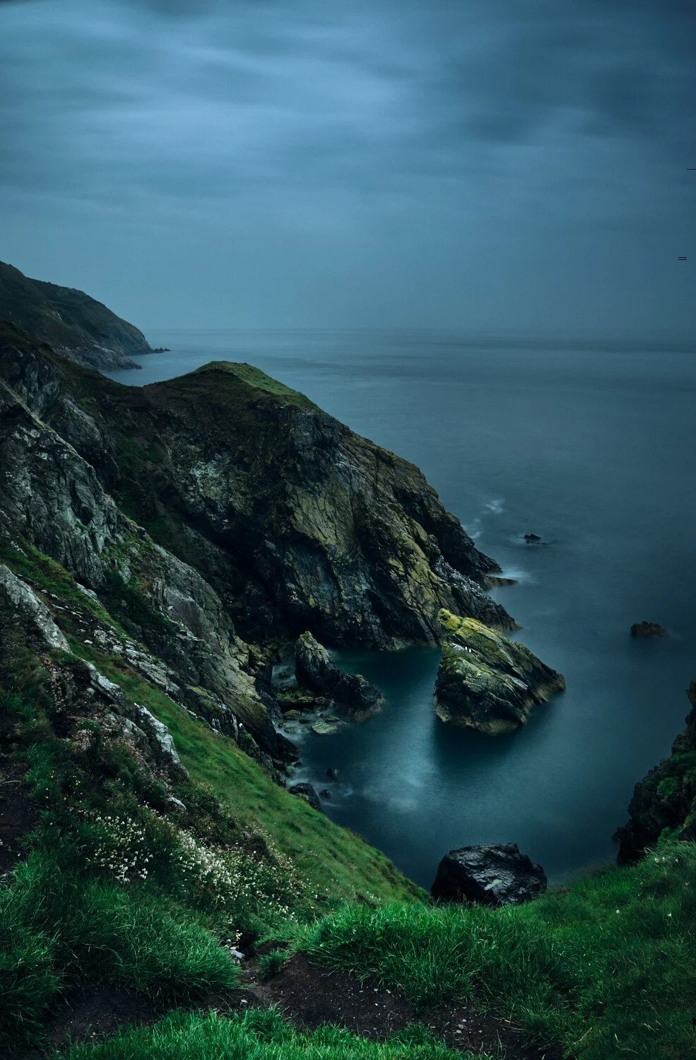Coastal cliffs with green grass and rocky shoreline, under a cloudy sky