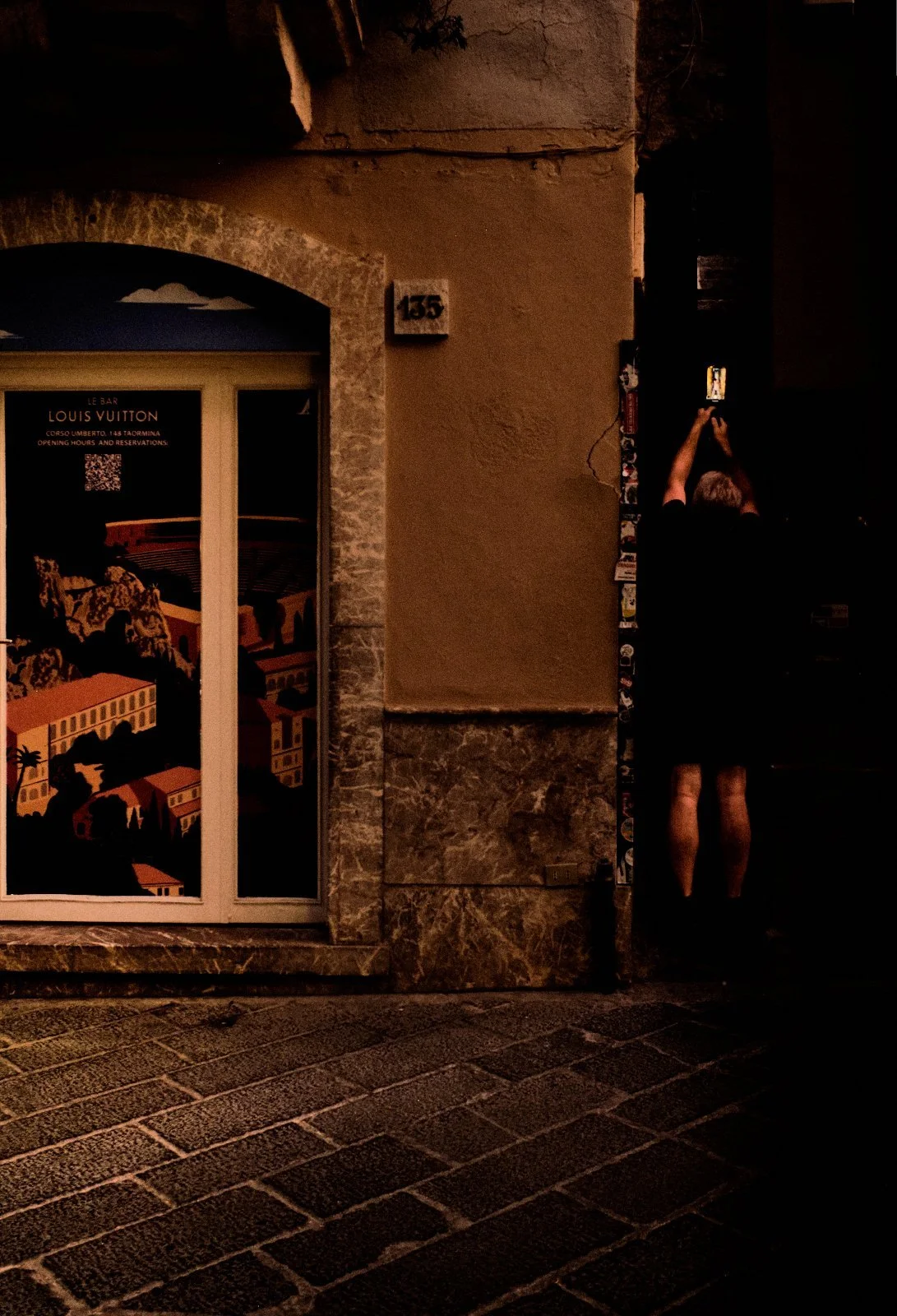 Man raising his phone in a dark doorway beside a luxury bar window illustrated with a stylised view of Taormina, Sicily — Provincia Malinconica by Tom Imio