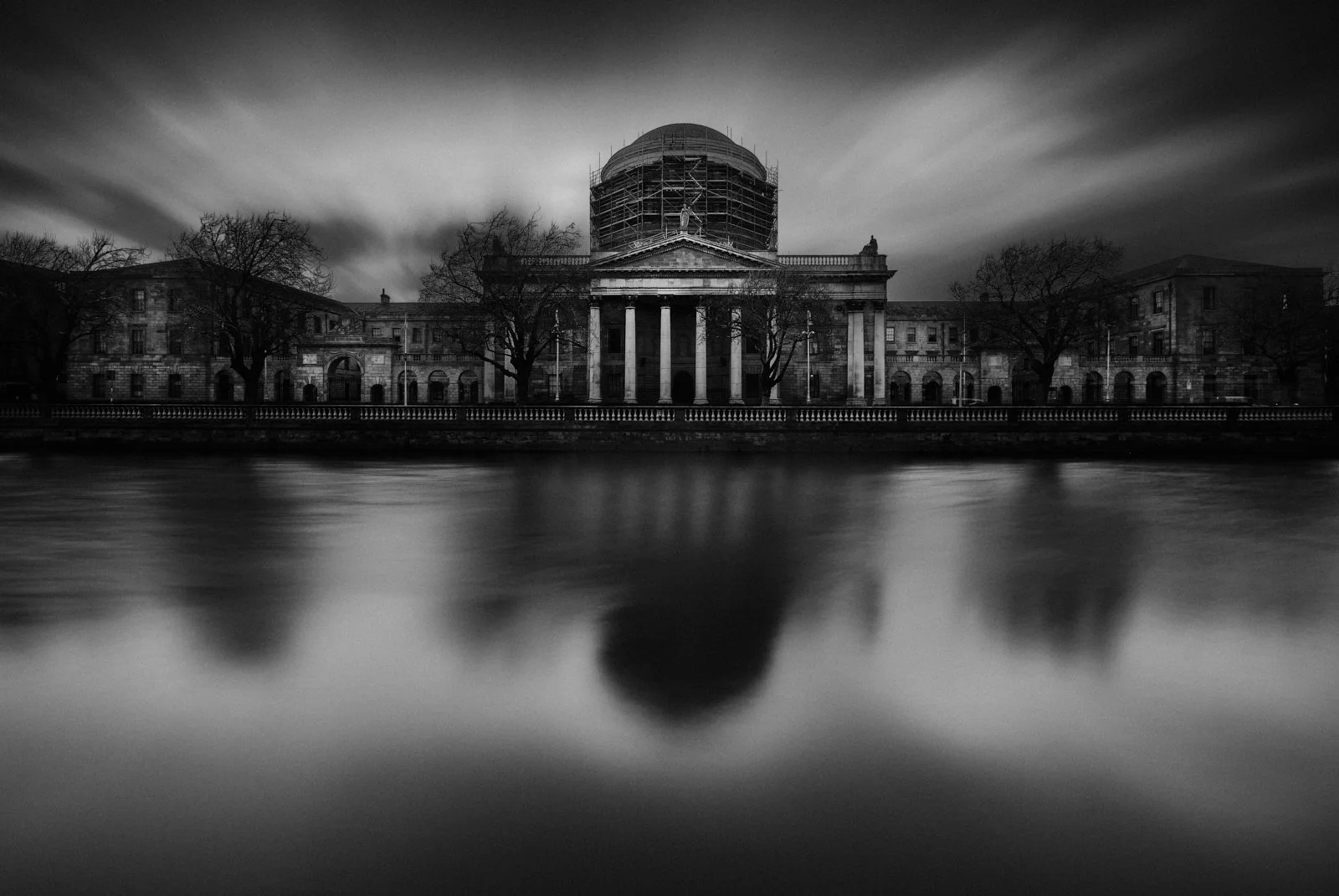 A black and white photo of a historic government building with classical architecture, trees, and scaffolding on the dome, reflected in a body of water in the foreground.