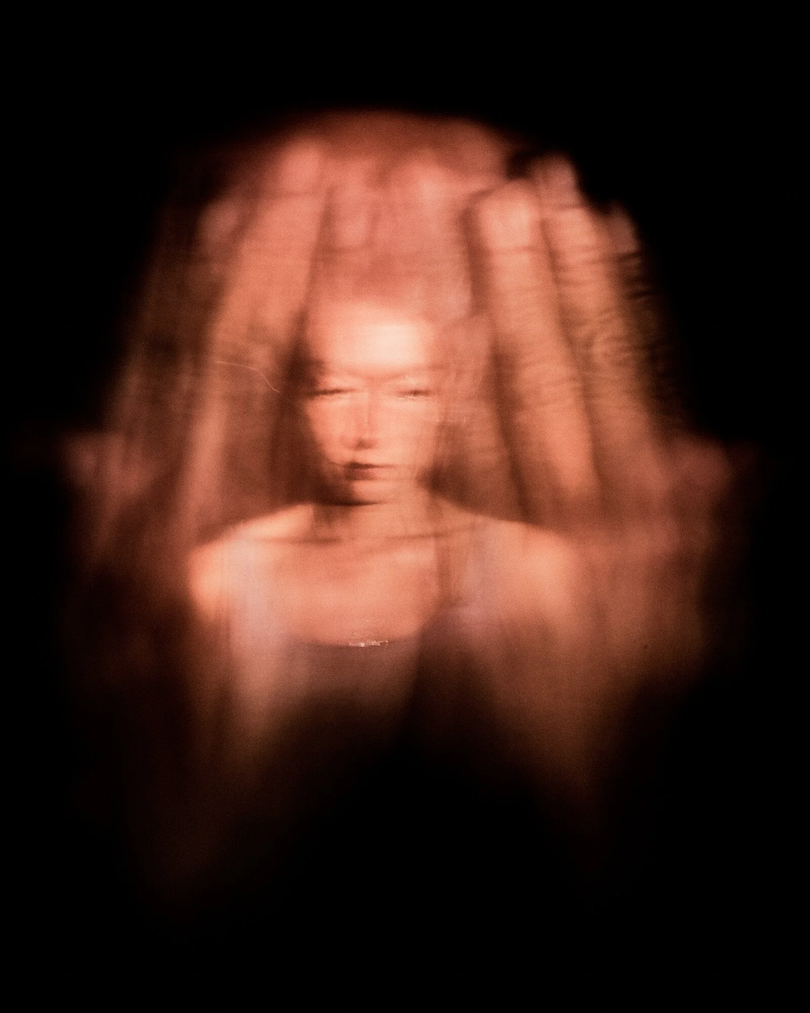 Blurred image of a woman with long hair and a serious expression, with her hands placed on her head, set against a dark background.