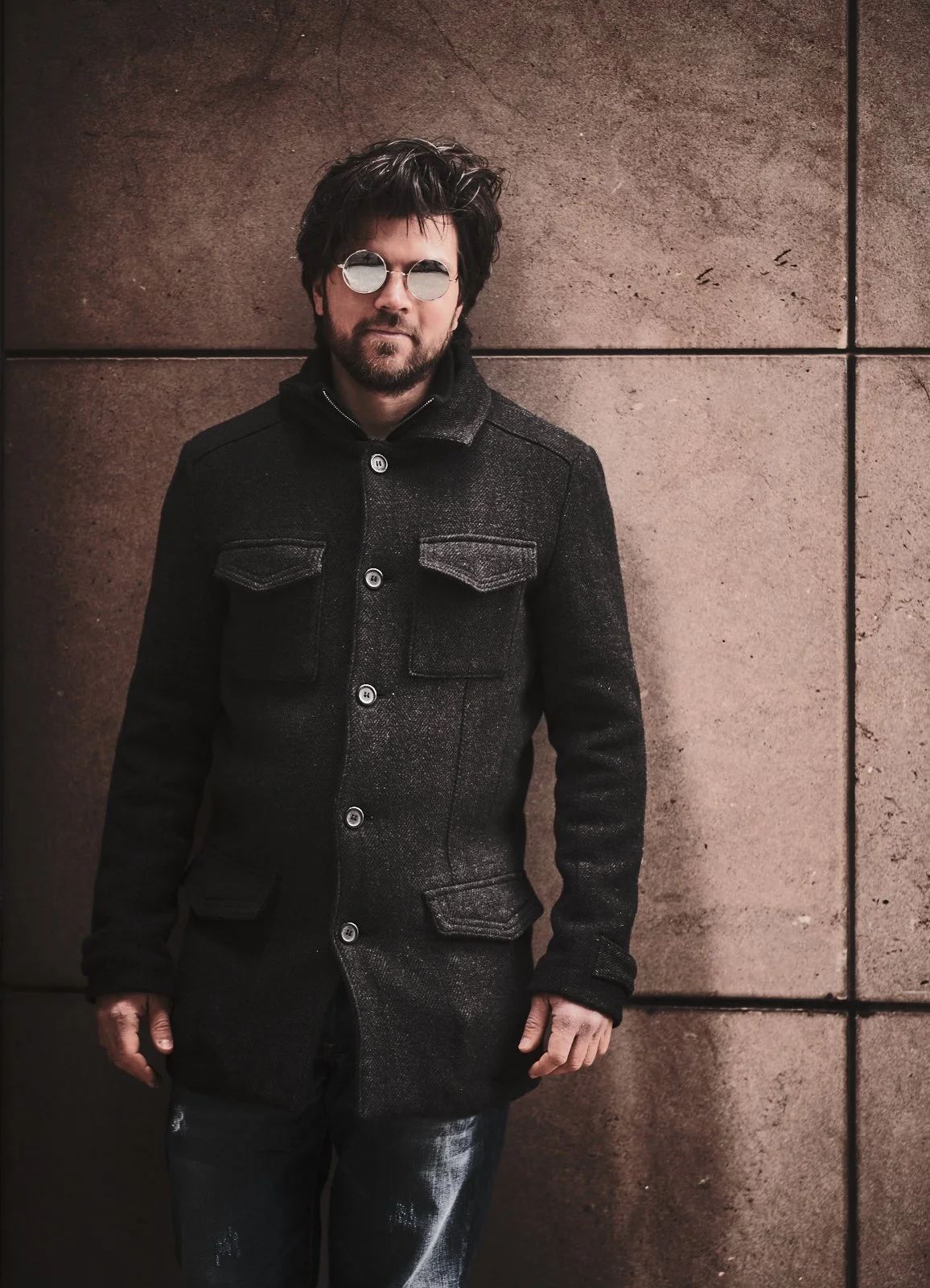 A man with dark, messy hair and a beard wearing round sunglasses and a black coat, standing against a tiled stone wall.