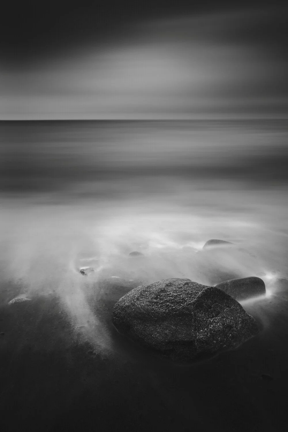 Black and white photo of a calm seashore with rocks in the foreground and blurred water and sky in the background.