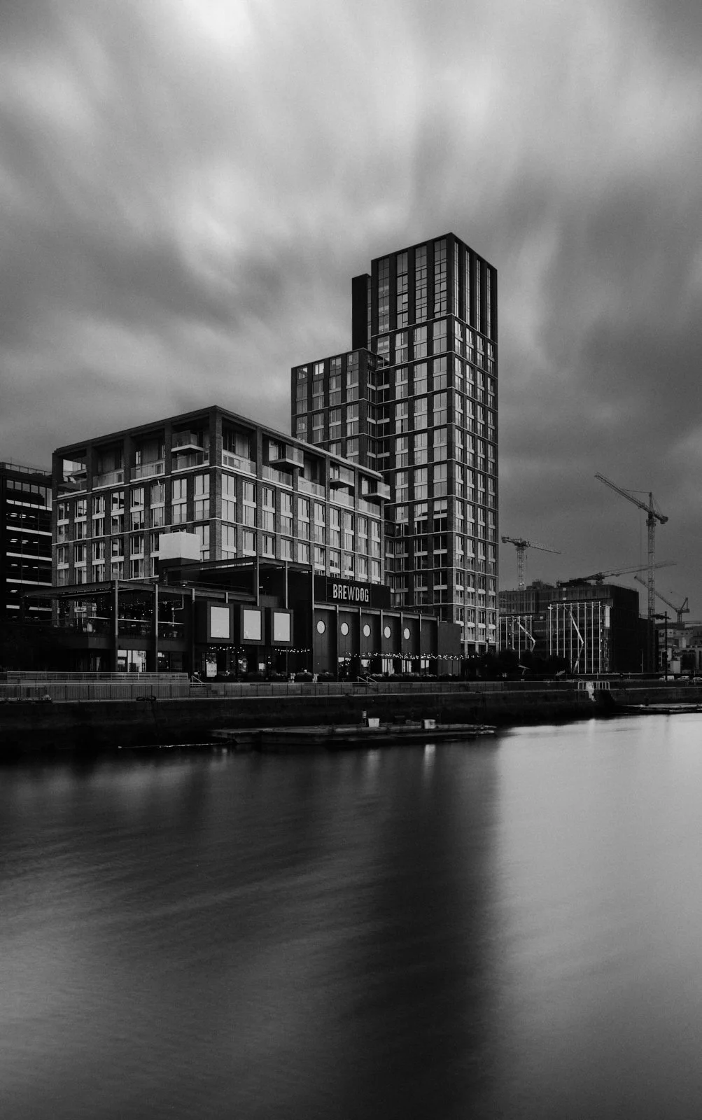 Black and white photo of modern buildings along a waterfront with a cloudy sky overhead.