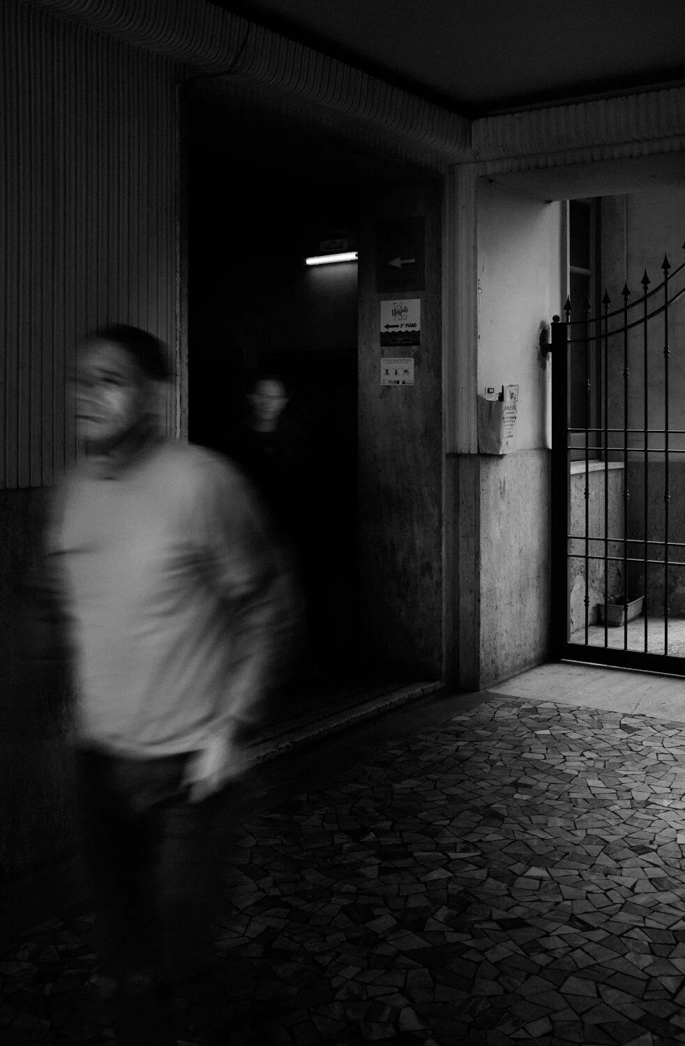 Blurry figure of a person walking past an open gate in a dark, urban environment at night.