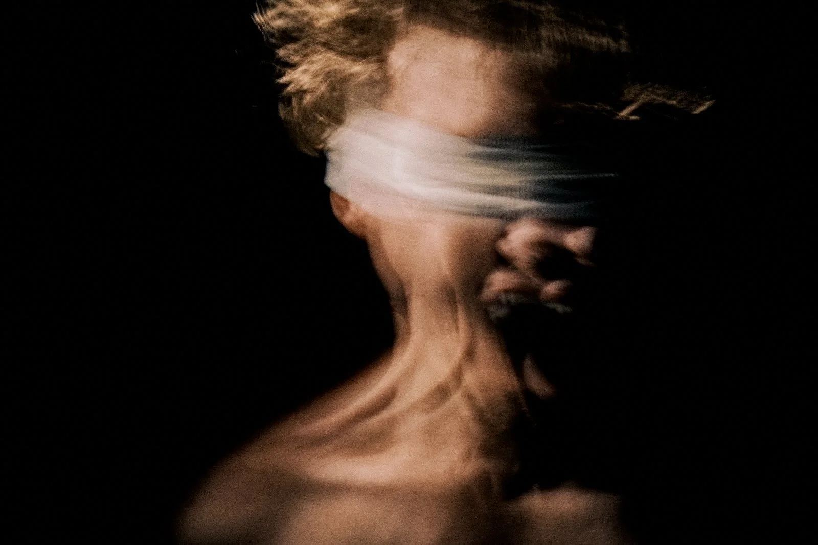 A person with short hair and a blindfold covering their eyes appears to be distressed, with their mouth open and neck strained, against a dark background.