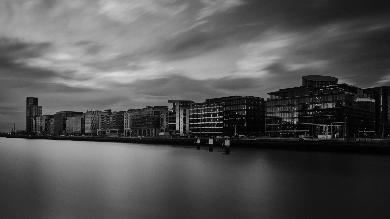 Modern cityscape with high-rise buildings along a waterfront, under a cloudy sky, in black and white.