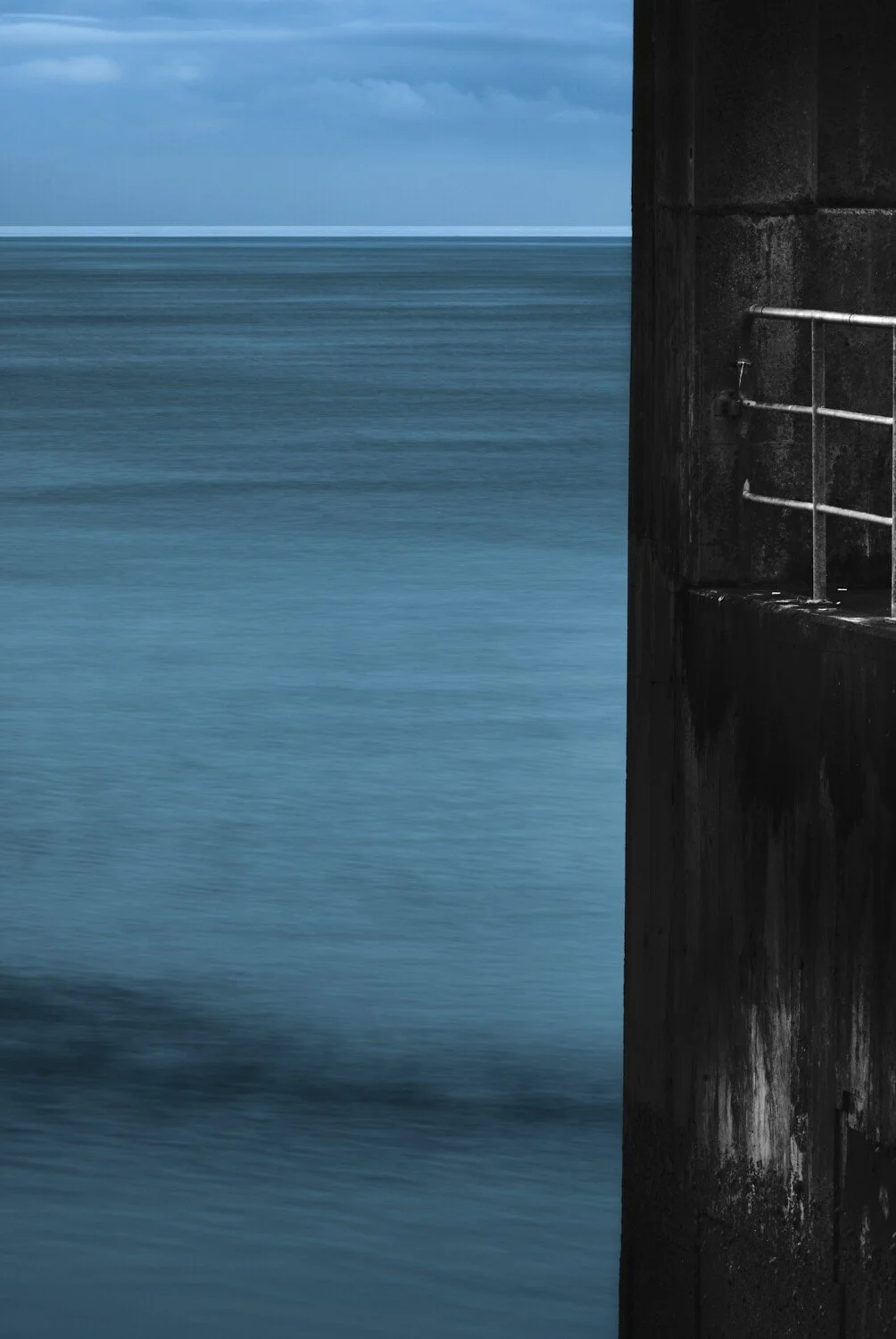 A black concrete structure with metal railing on the right side, overlooking a calm blue ocean under a cloudy sky.