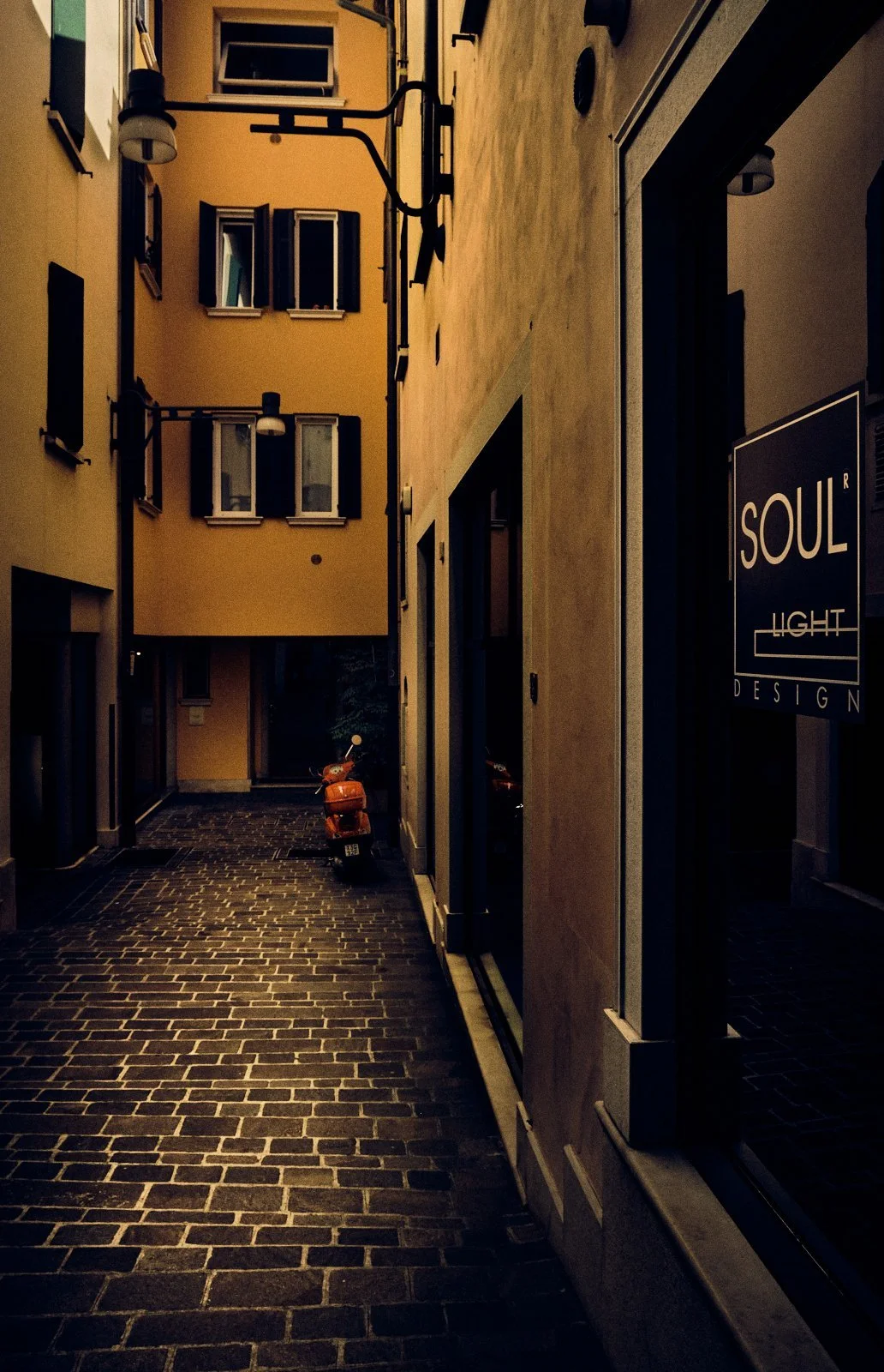 Illuminated design shop sign in a deserted cobbled alley with an orange scooter, Desenzano del Garda, Italy — Provincia Malinconica by Tom Imio