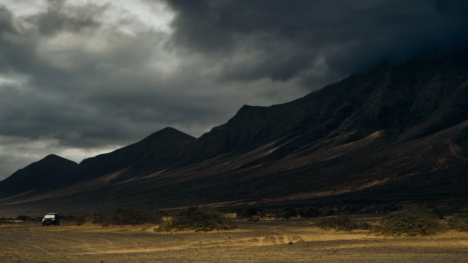 A dark, cloudy sky over a mountain landscape with a sandy, open plain and a single vehicle in the distance.