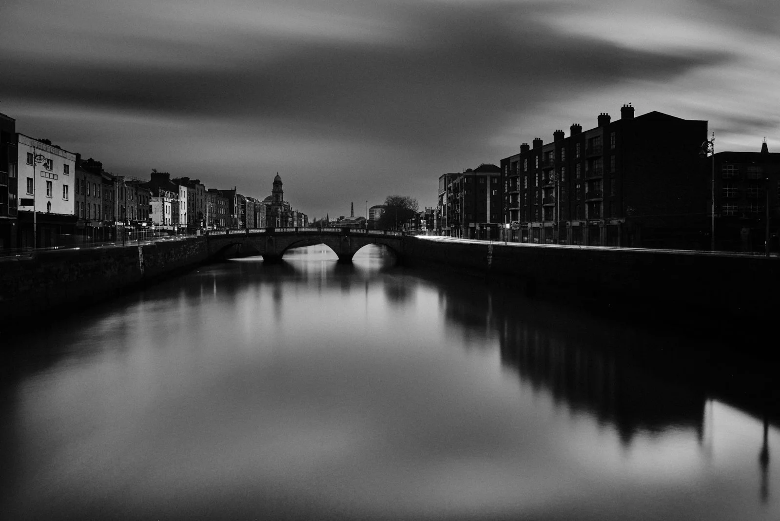 A black and white photo of a cityscape taken from across a river, with a bridge and buildings on both sides, under a cloudy sky.