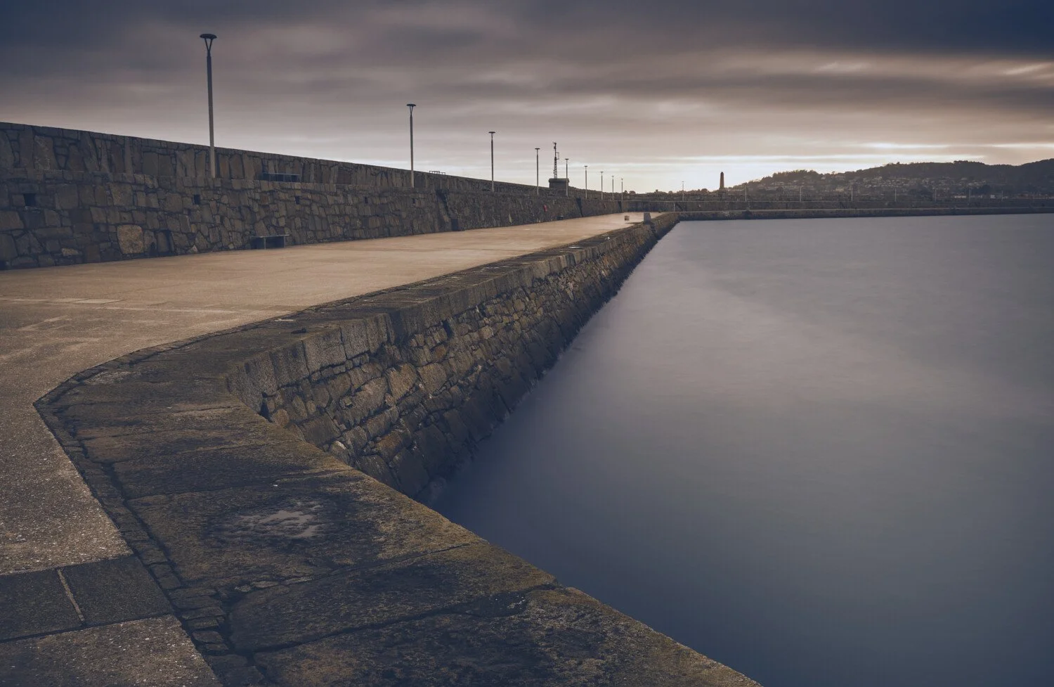 Empty stone pier with railing and streetlights, calm water, cloudy sky, distant hilly landscape.