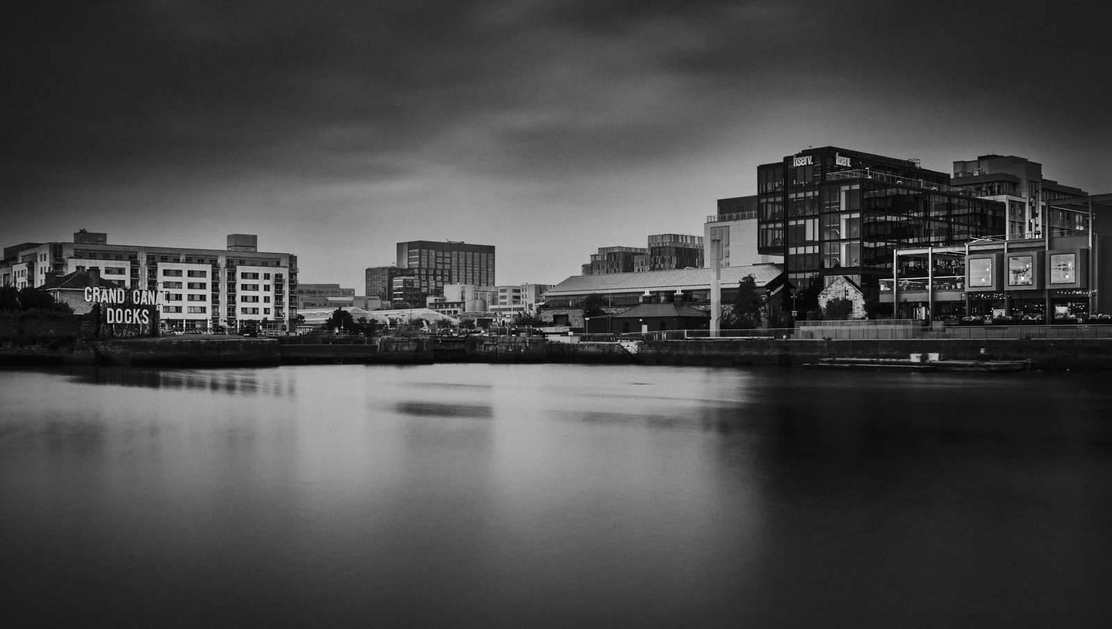 Black and white photo of an urban waterfront scene with modern buildings, a sign reading 'Grand Canal Docks,' and calm water reflecting the cityscape.