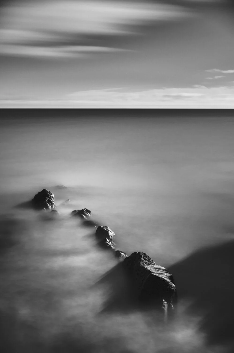 Black and white photo of rocks in the water with calm ocean and cloudy sky in background.