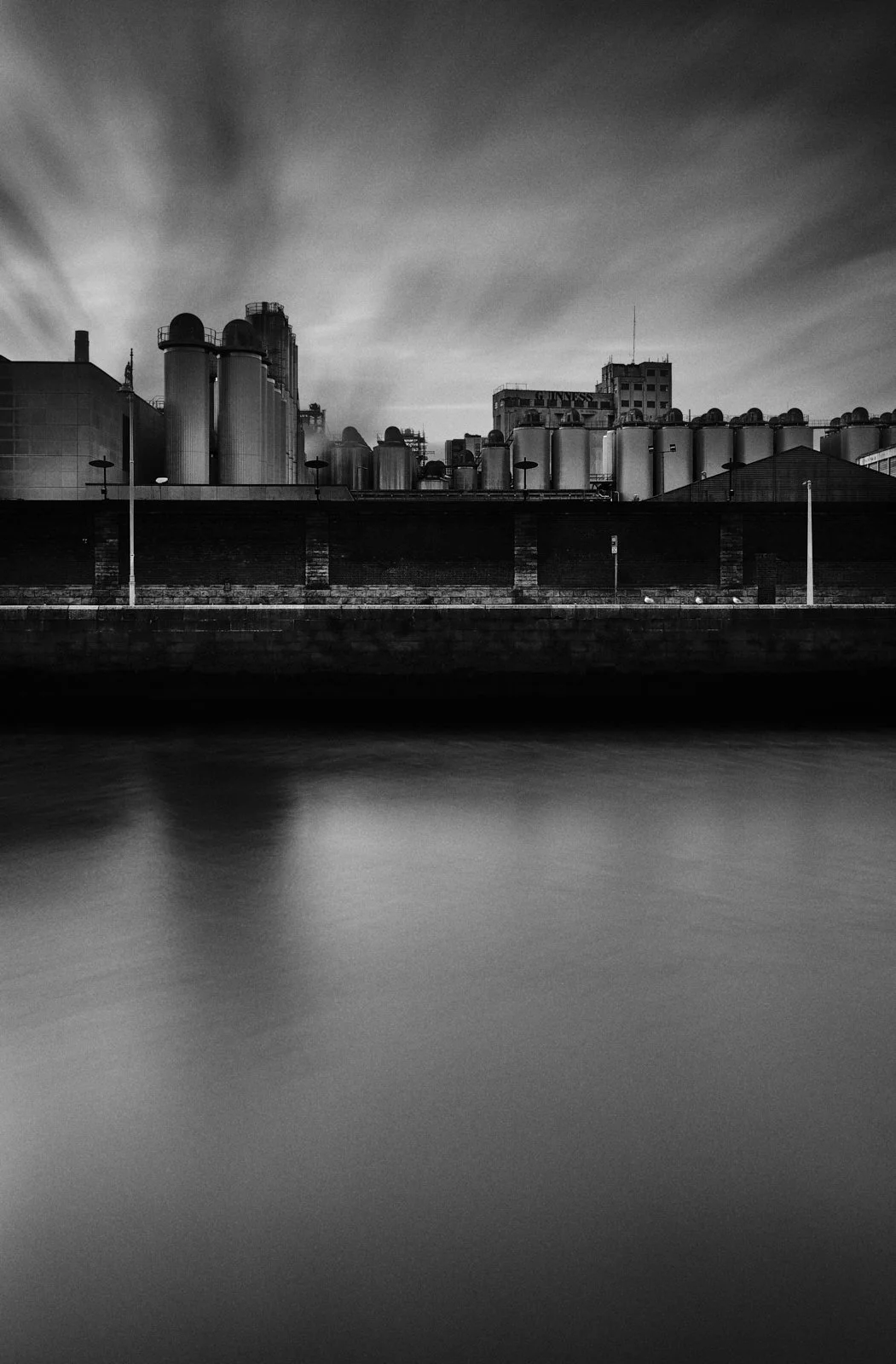 Black and white photo of an industrial building with large silos next to a body of water, with a cloudy sky.