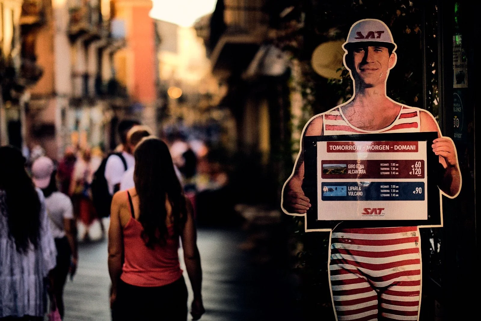 Faded cardboard figure in a striped bathing suit advertising Etna tours as tourists stroll past in Taormina, Sicily — Provincia Malinconica by Tom Imio