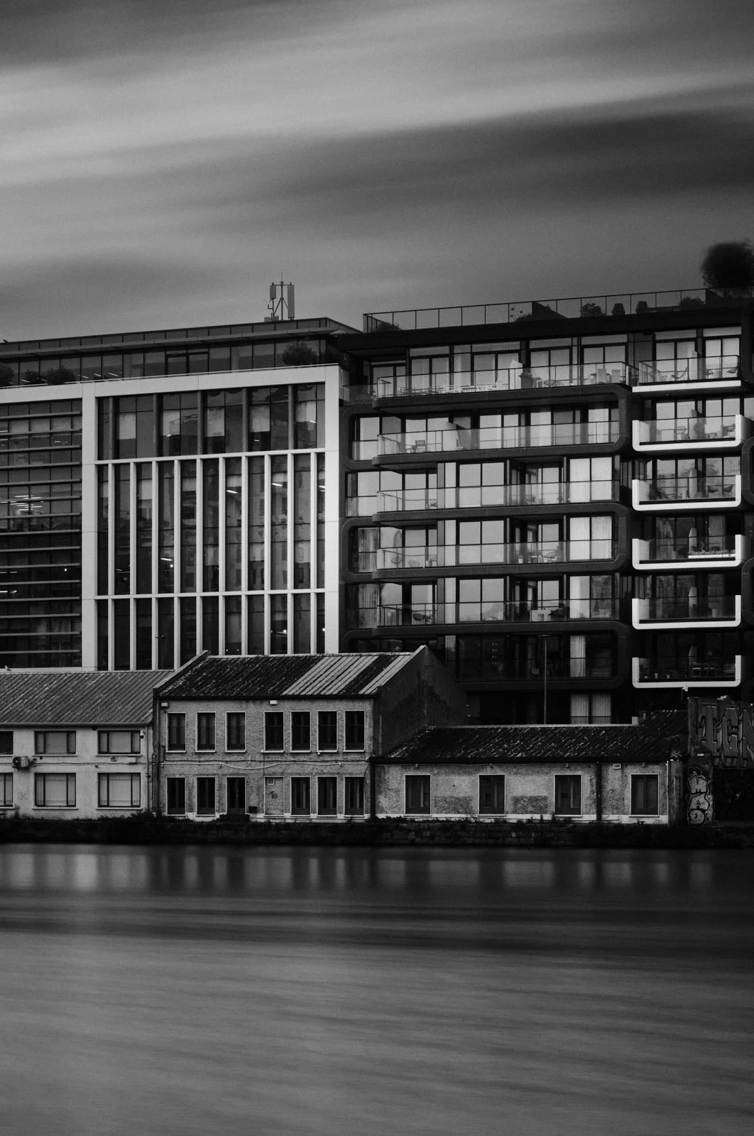 Black and white photo of modern apartment buildings and an older small house along a river with a cloudy sky.