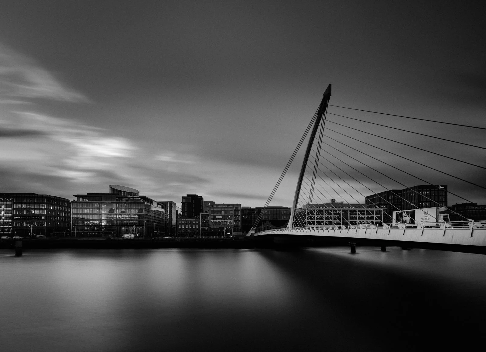 Nighttime view of a city skyline with modern buildings, a bridge with a tall, angled pylon and cable supports, and smooth water in the foreground.