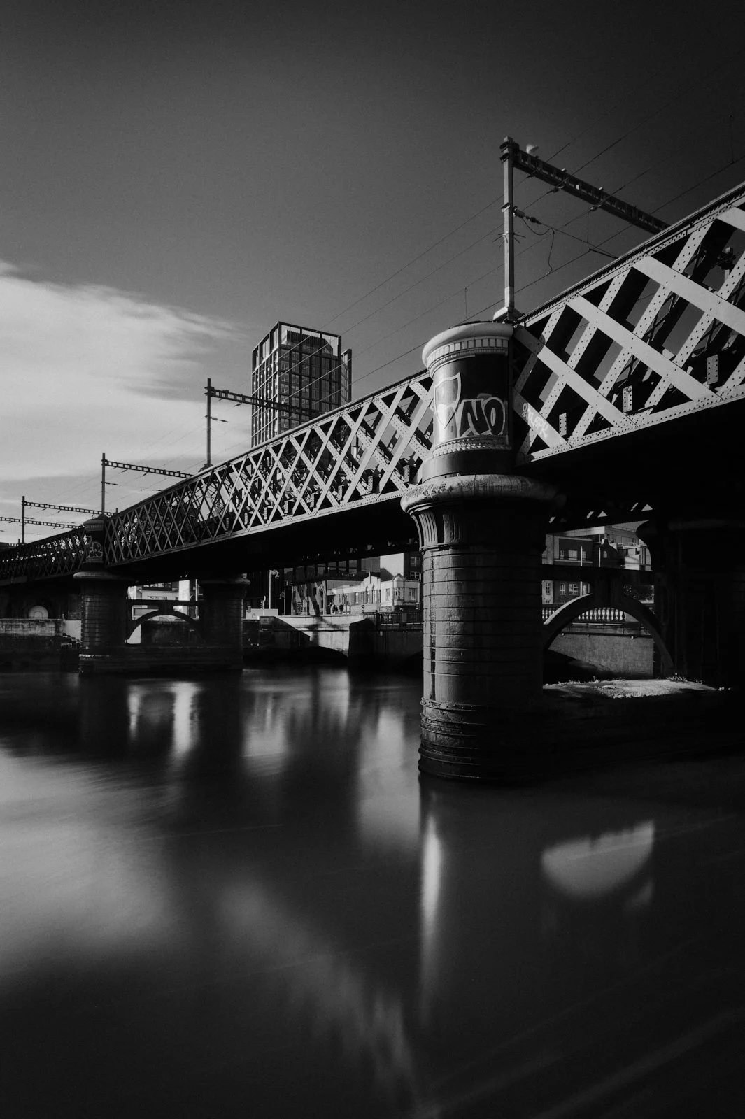 Black and white photo of a city bridge over a river, with a tall modern building in the background.