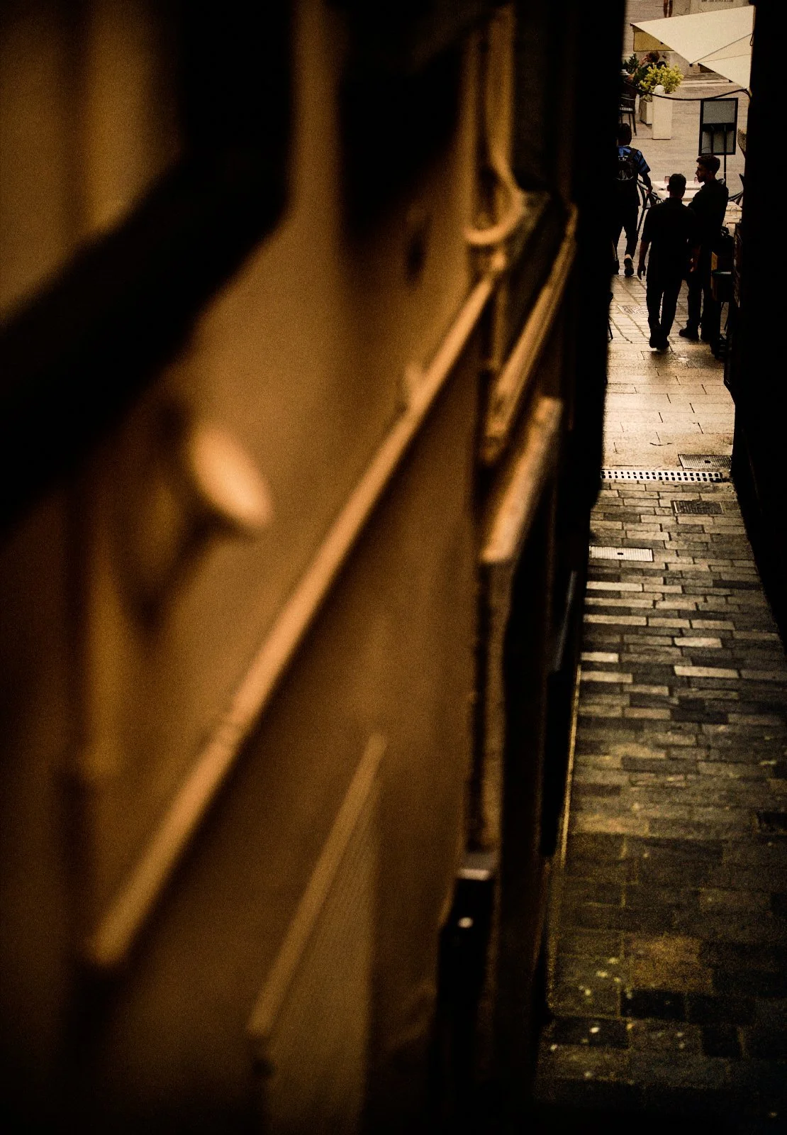 View from a dark narrow alley onto a sunlit square with passersby and café umbrellas in Desenzano del Garda — Provincia Malinconica by Tom Imio