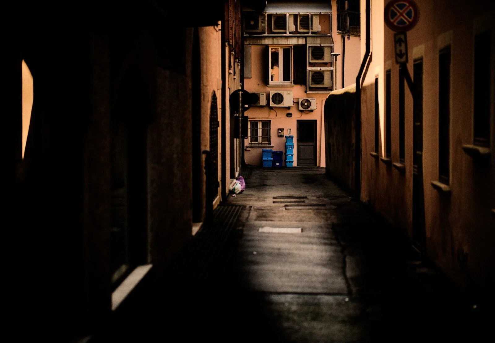 Air conditioning units and plastic crates on a faded pink facade at the end of a shaded alley in Desenzano del Garda — Provincia Malinconica by Tom Imio