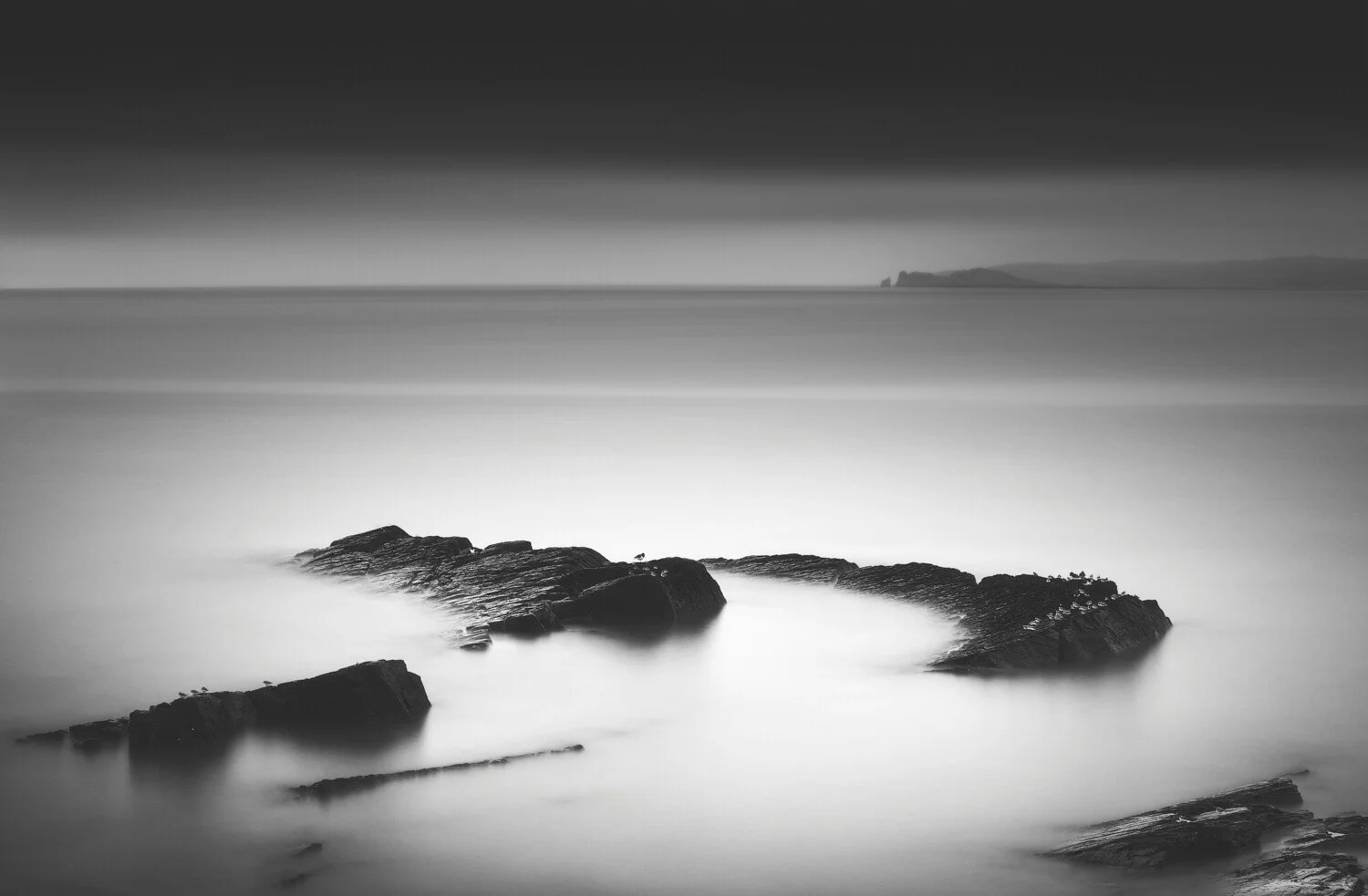 Black and white photo of rocks in water with a distant landmass on the horizon