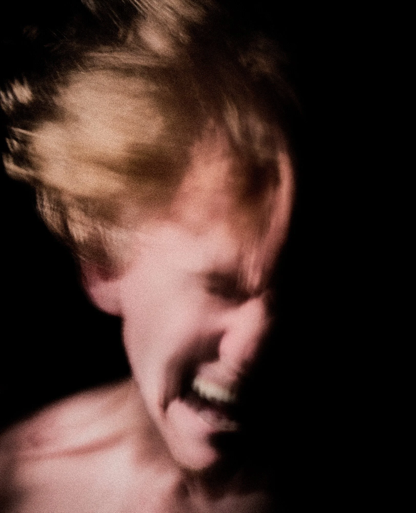 Close-up of a person with short hair laughing or smiling with their eyes closed, against a dark background.