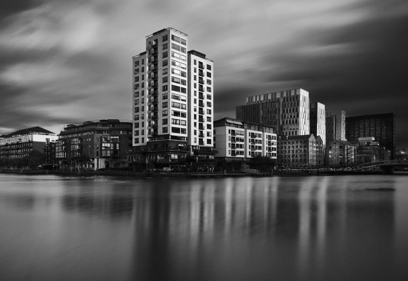 Black and white photo of modern high-rise buildings along a river with a blurred water surface and cloudy sky