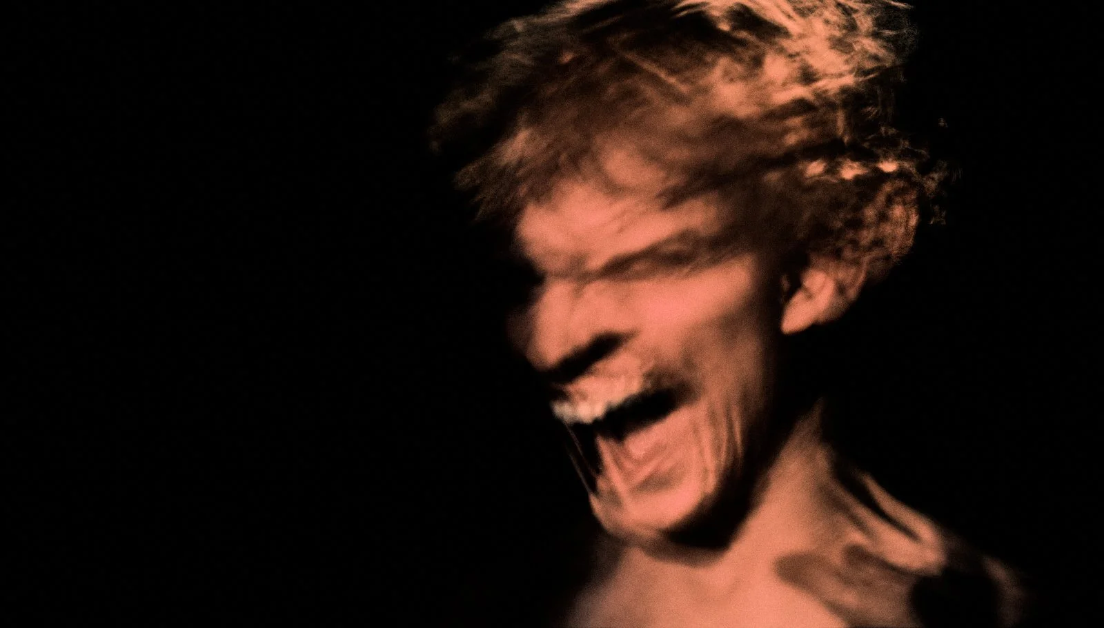 A close-up of a person's face with a blurred, motion effect, smiling or laughing, with tousled hair and a dark background.