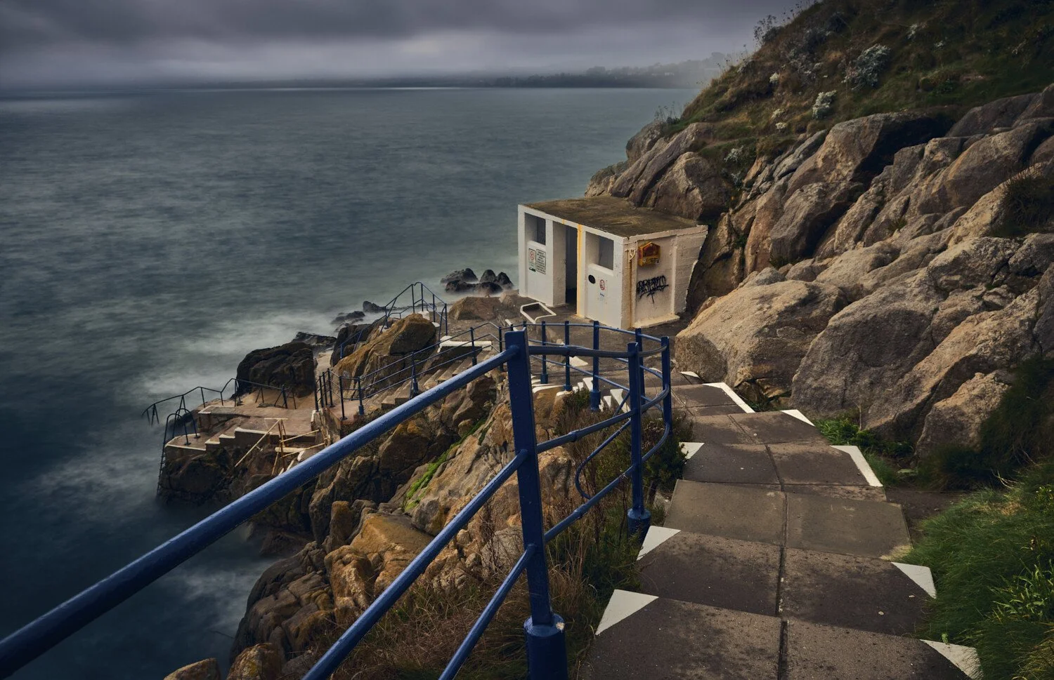 A rocky coastline with a winding concrete pathway, blue rails, and a small white utility building near the water, under cloudy skies.