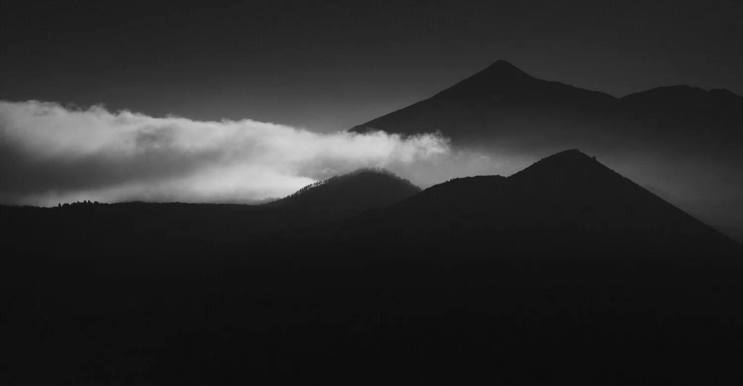 Black and white landscape of mountain silhouettes with a prominent peak and a layer of clouds in the sky.