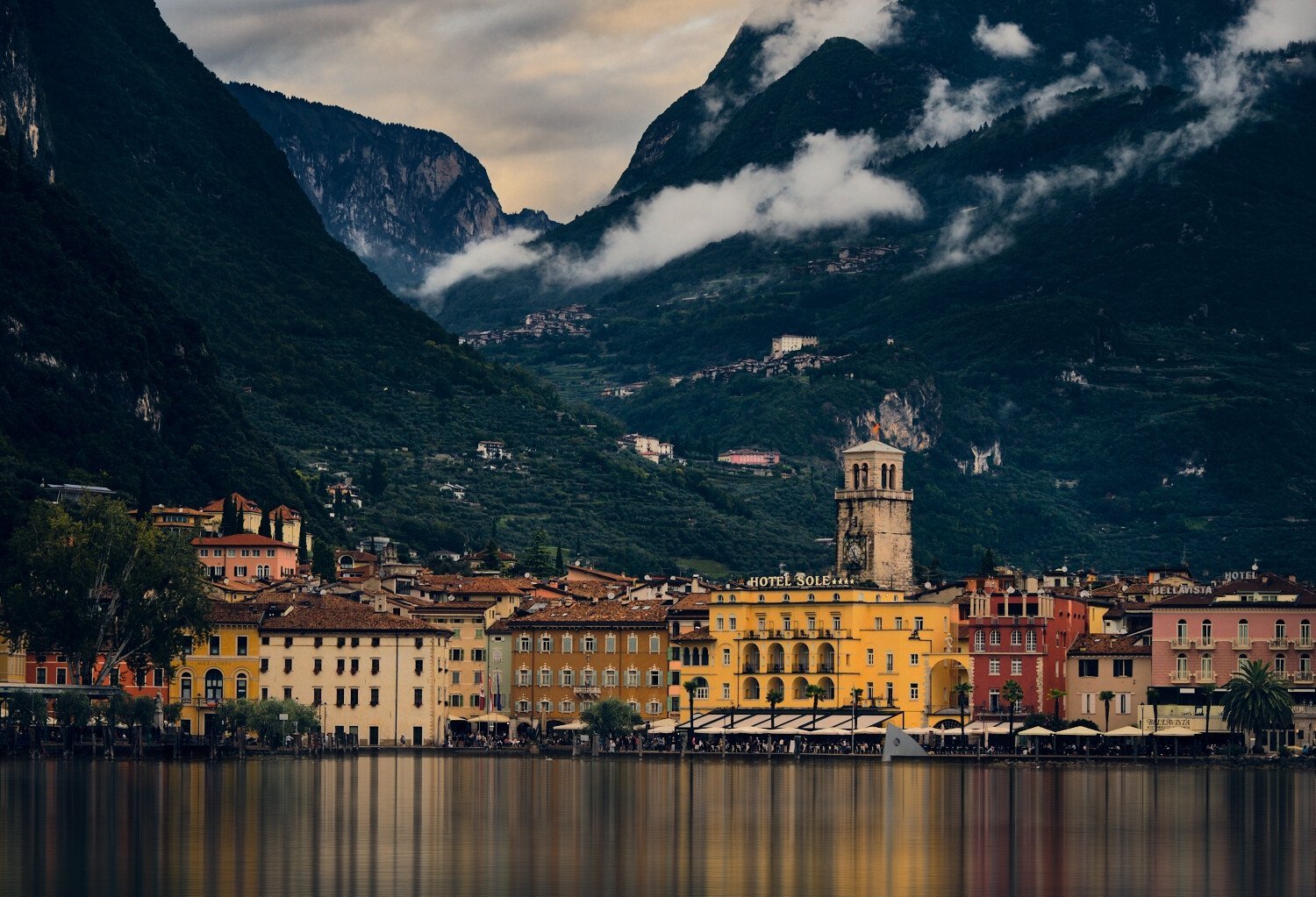 Colorful buildings along a waterfront with a mountain backdrop, including a clock tower and lush green hills partially covered with clouds.
