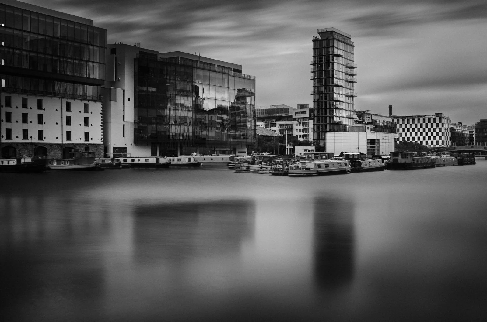 Black and white photo of modern buildings along a waterway with boats docked at the edge, under a cloudy sky.