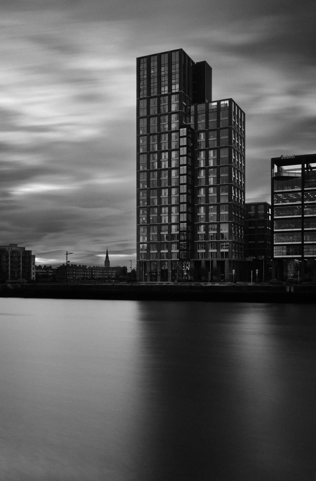 Black and white photo of modern high-rise building near water, with clouds in the sky.