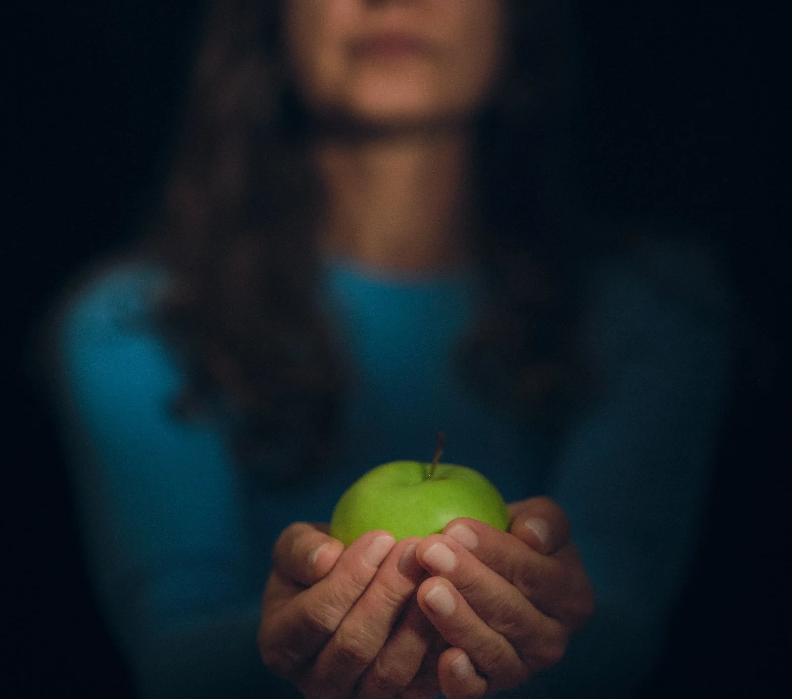 A person holding a green apple with both hands, with a blurred person in the background. Jabłko Adama