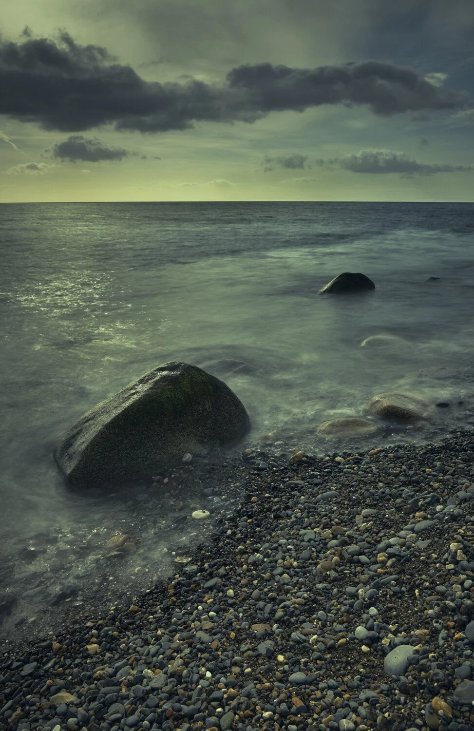 A rocky seashore at dusk with dark clouds overhead and calm ocean water.